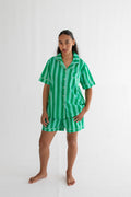 A woman modeling a two-piece green striped pajama set, consisting of a short-sleeve button-up shirt and matching shorts, against a plain white studio background.