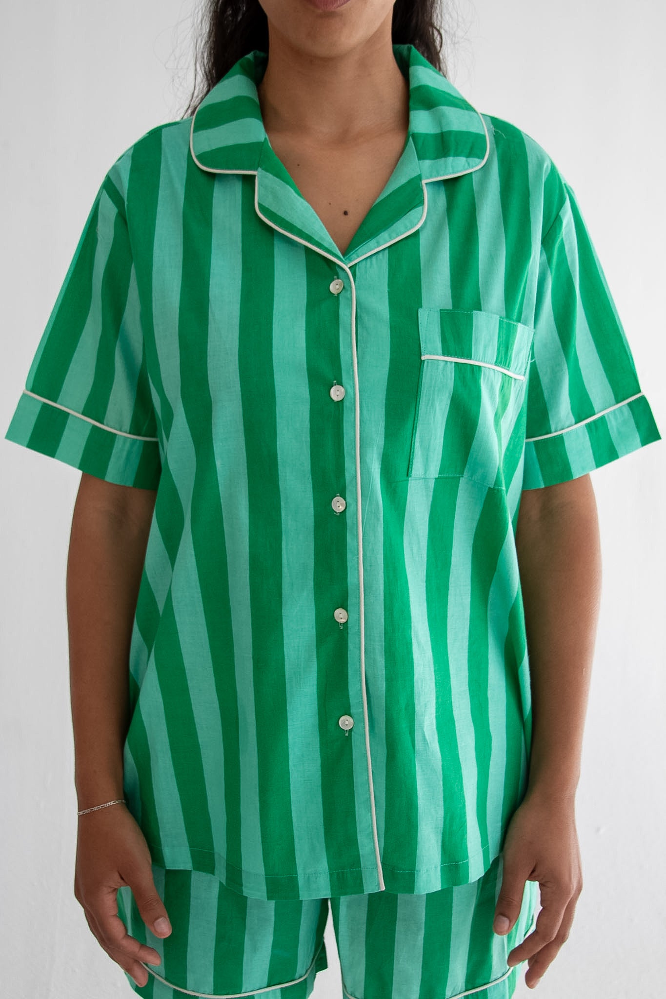A person wearing a green and mint striped short-sleeve pajama shirt with white piping, paired with matching striped pajama shorts, shown against a plain light background.